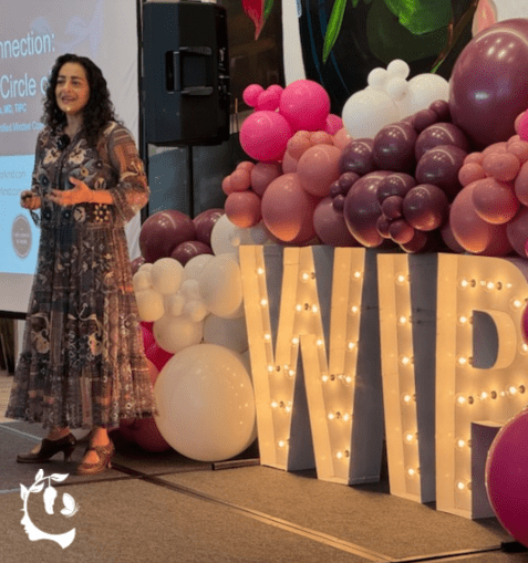 A woman in a floral dress speaking on stage at a WIP (Women in Psychiatry/Professional) event, standing beside large illuminated marquee letters "WIP" surrounded by a pink, mauve, and white balloon arch.