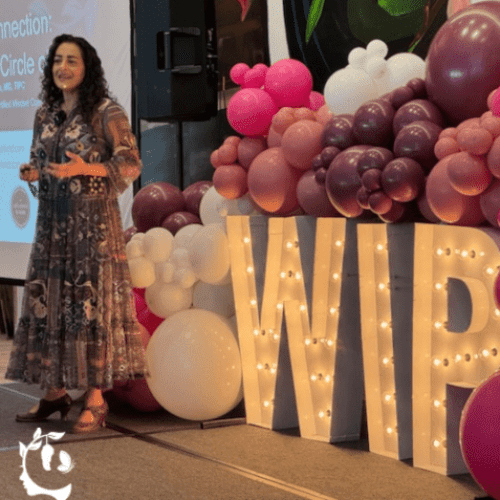 A woman in a floral dress speaking on stage at a WIP (Women in Psychiatry/Professional) event, standing beside large illuminated marquee letters "WIP" surrounded by a pink, mauve, and white balloon arch.